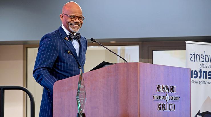 James Ray Rhodes standing at a podium in a suit with his award places in the foreground